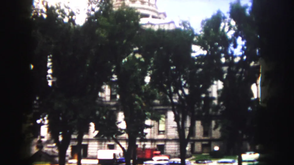 Denver Colorado State Capitol appears behind dark tree trunks and foliage in 1958 8mm footage, with small cars near the base.
