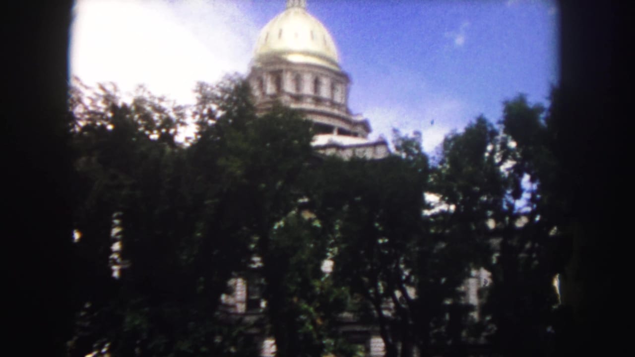 1958 Denver home-movie frame showing the Colorado State Capitol dome partly obscured by trees, with dark 8mm frame edges.