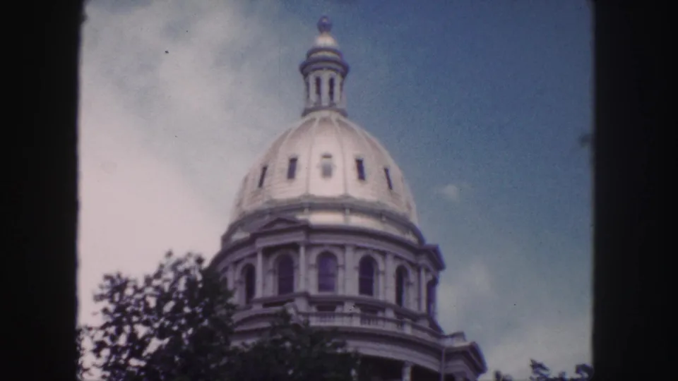 1958 Denver 8mm frame showing the Colorado State Capitol dome from below, with trees at the base and soft color film grain.