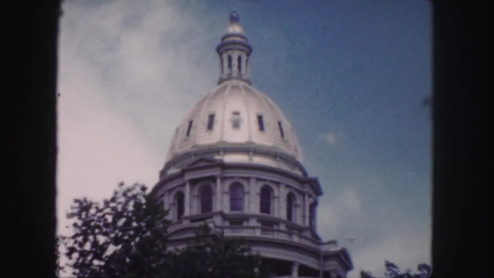 1958 Denver home-movie still of the Colorado State Capitol dome and upper facade, framed by leafy trees and visible film borders.