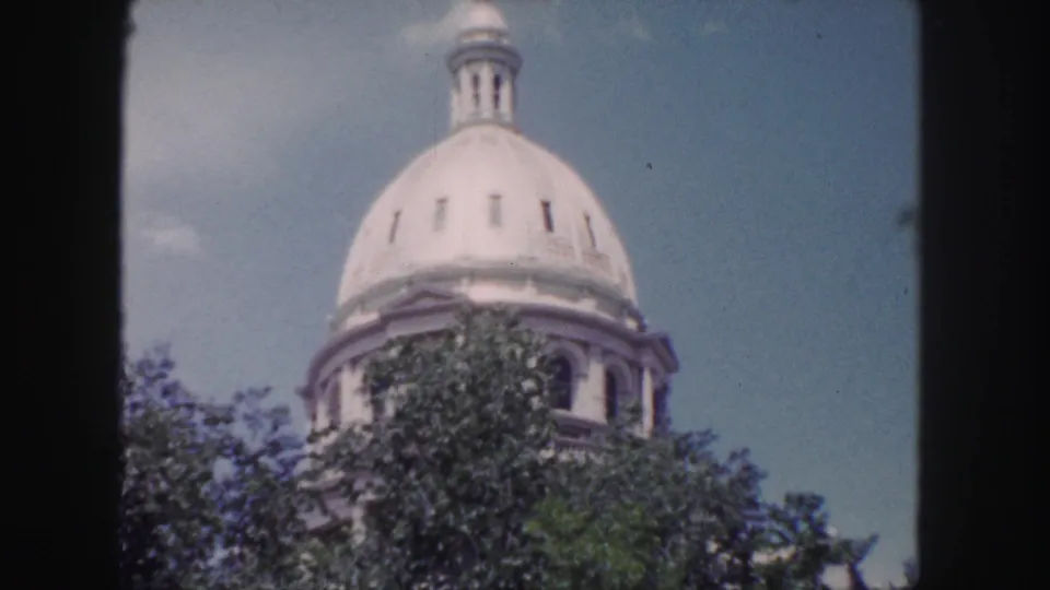 Wider 1958 Denver frame showing the white dome and cupola above green trees, with soft color and visible 8mm grain.