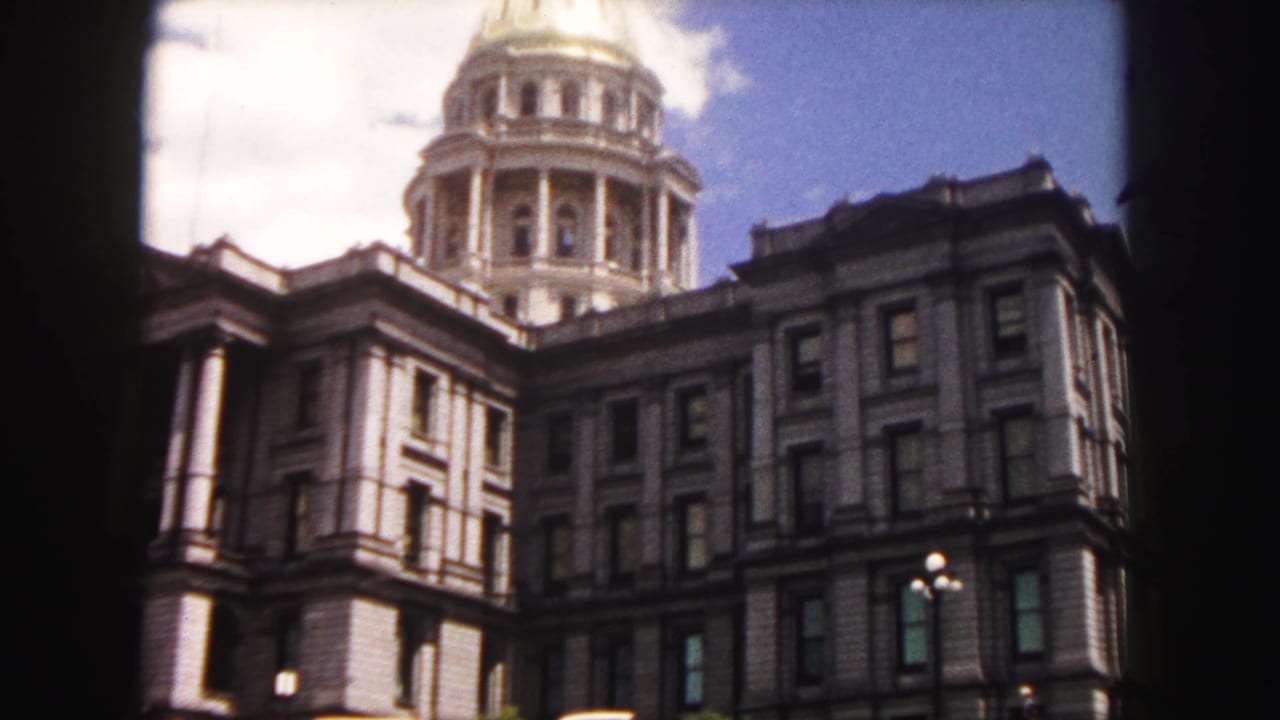 Capitol building exterior with dome, columns, dark film border, sky, and cars along the curb.