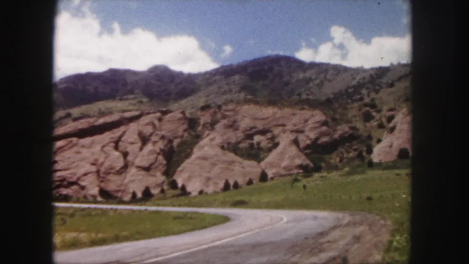 1958 Denver-area Red Rocks Park view with a curving road, red rock formations, green grass, mountains, and scattered clouds.
