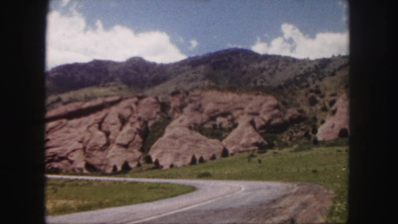Curving road beside red rock formations, green grass, hills, and blue sky.