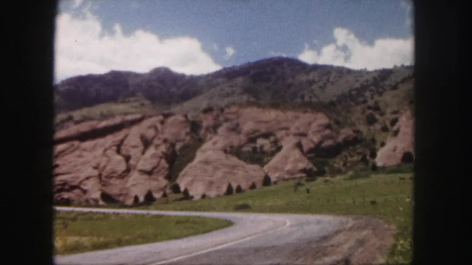 1958 8mm view near Denver of a winding road passing red rock formations below grassy hills and clouds.