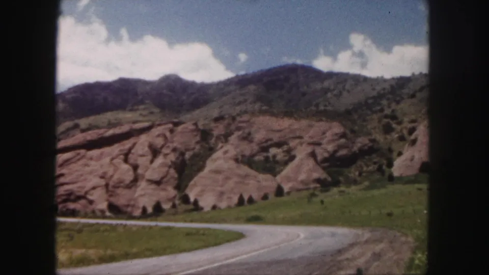 A framed 8mm shot of red rock formations, green roadside grass, and a curving road near Denver, Colorado.