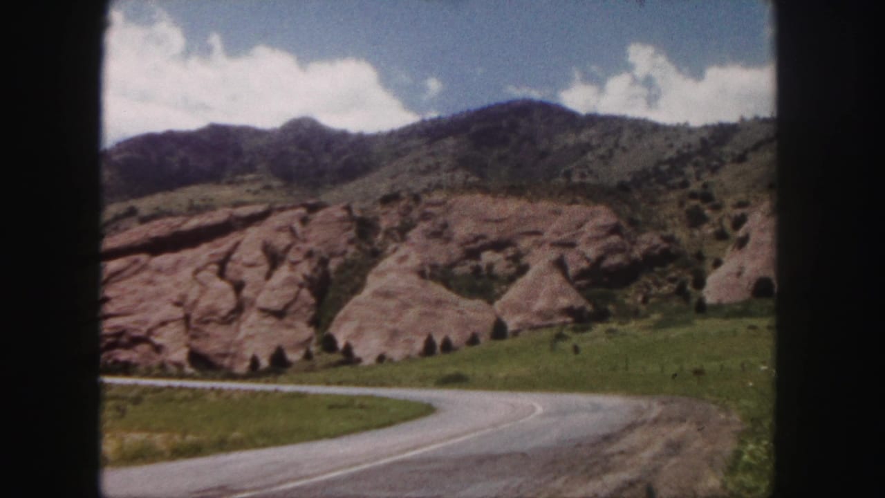 1958 8mm view near Denver showing a curved paved road passing grassy slopes and reddish rock formations beneath distant foothills.