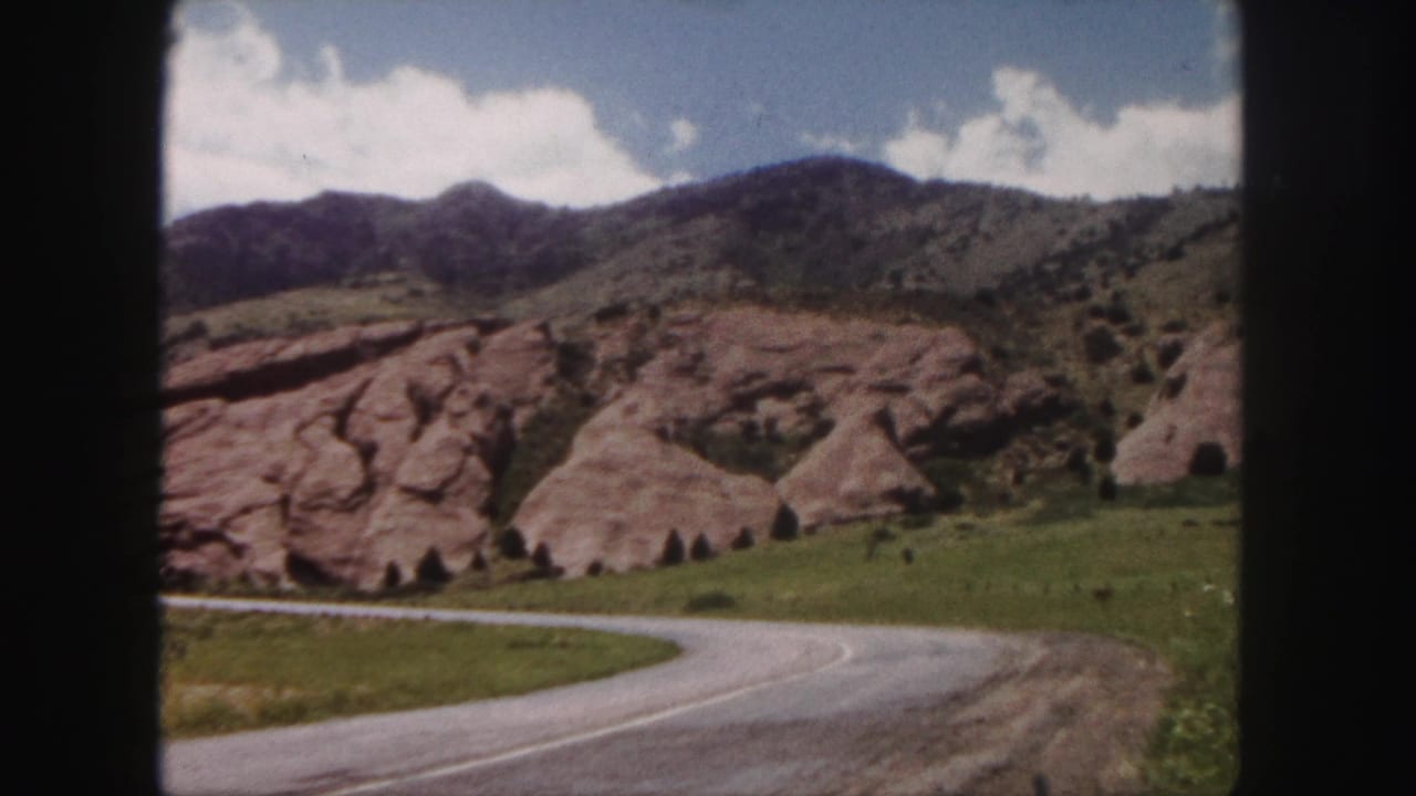 Curving road, red rock outcrops, green hillside, mountains, and cloudy sky.