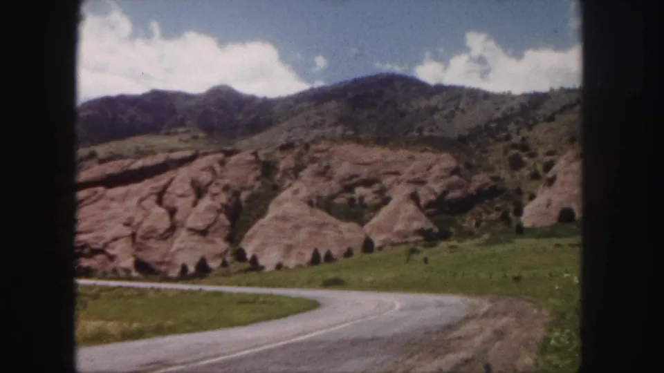 1958 Colorado home-movie frame of a road curving past red rock outcrops and green foothills under a bright cloudy sky.