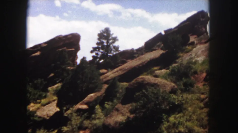 1958 Colorado 8mm frame showing reddish roadside rock formations, low shrubs, and a single pine tree against a bright sky.