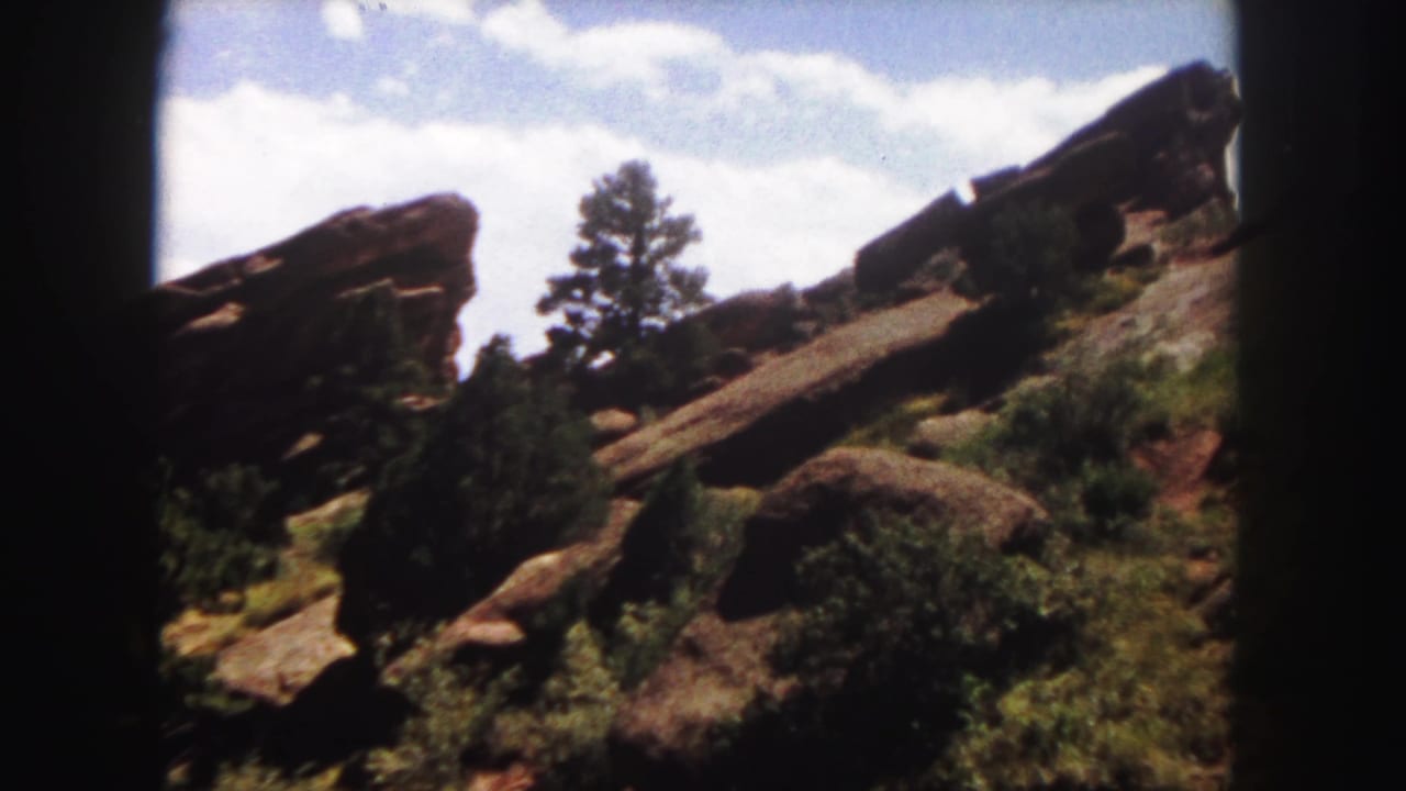 1958 Denver-area 8mm frame showing red sandstone outcrops, evergreen trees, shrubs, clouds, and dark film-gate edges.