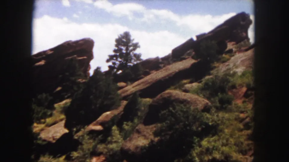 1958 Denver-area 8mm frame showing a red rock formation, a tall evergreen, shrubs, and soft clouds above the slope.