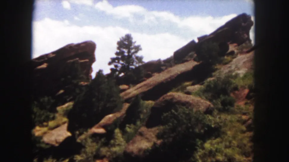 1958 Denver-area frame with red sandstone ledges, dark evergreens, scrub vegetation, and visible 8mm frame edges.