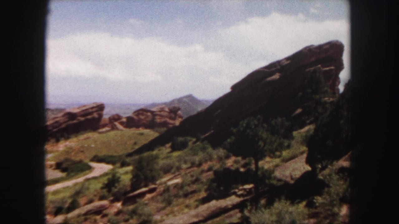 Red rock formation on a green hillside with shrubs, a winding path, distant ridge, cloudy sky, and dark film gate edges.