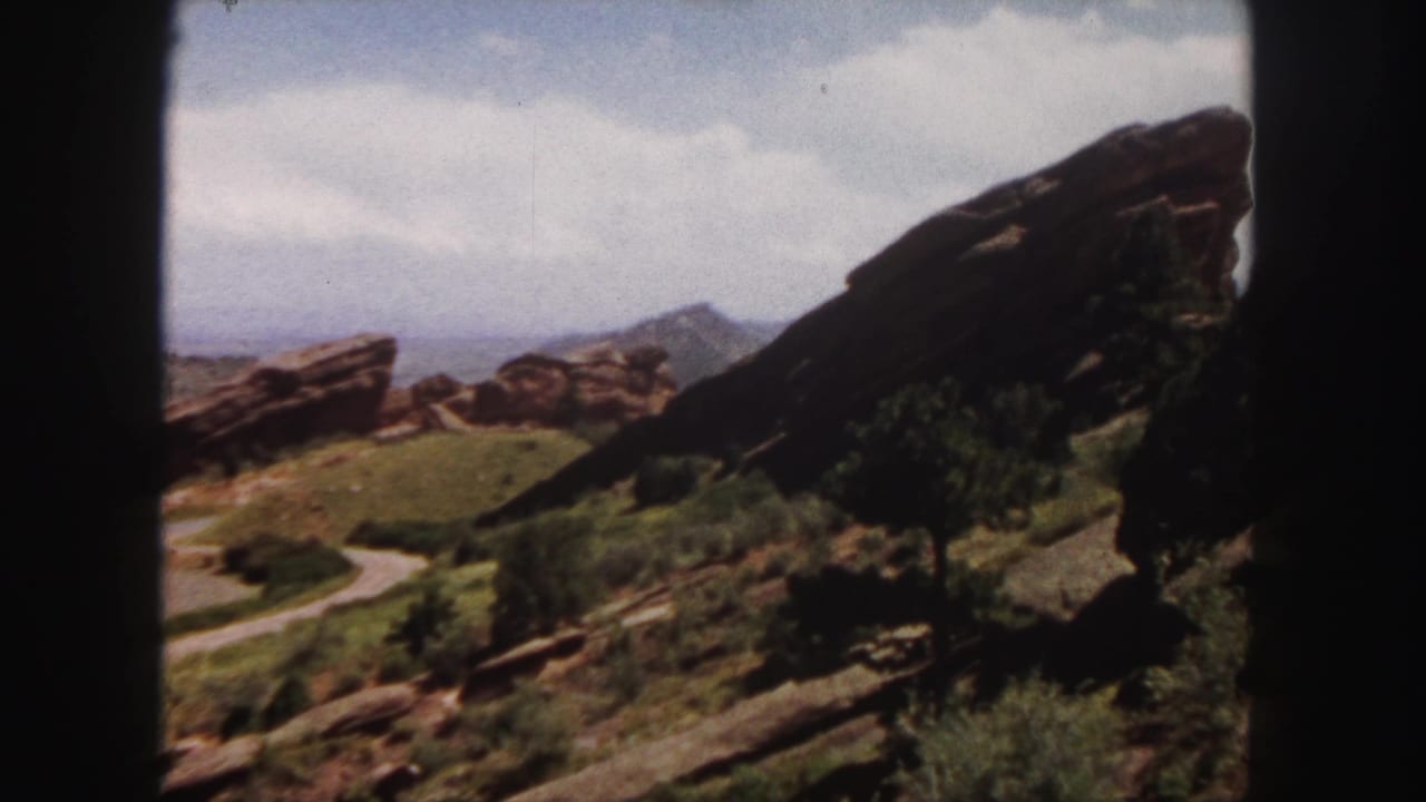 Sandstone cliffs and boulders overlook a green slope with shrubs, a curved path, distant mountains, and cloudy sky.