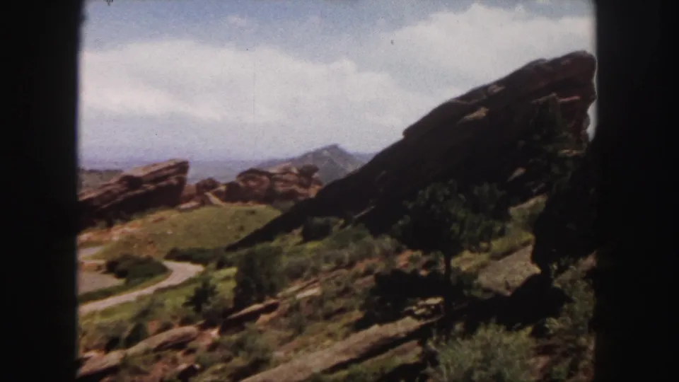 Denver-area 1958 home-movie frame with jagged red rocks, hillside vegetation, a curving path, distant ridges, and dark projector-gate borders.