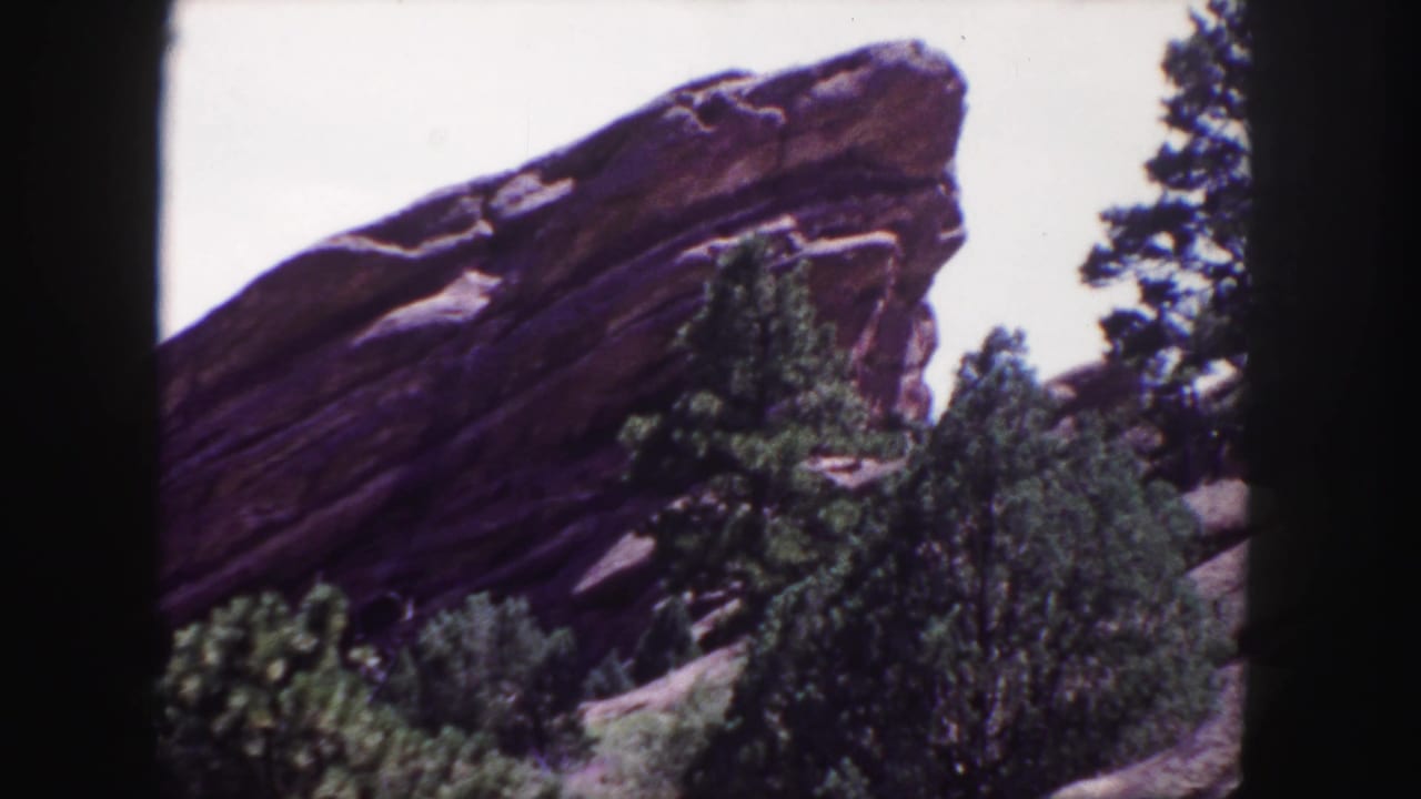 Large angled sandstone formation rises behind evergreen trees and shrubs with pale sky above.
