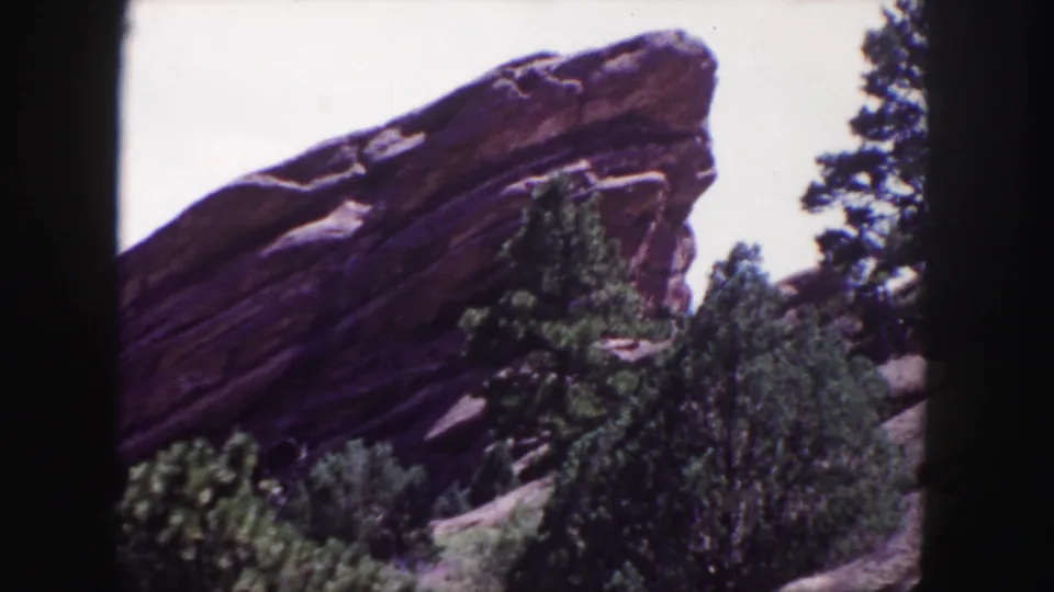 Close 1958 8mm view at Red Rocks near Denver, with a massive tilted sandstone slab partly screened by evergreens and brush.