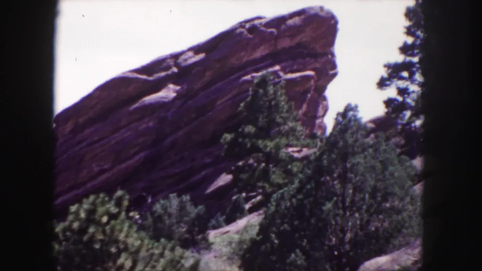 1958 home-movie frame of Red Rocks near Denver, focused on a sloping sandstone face, evergreen trees, and foreground brush.