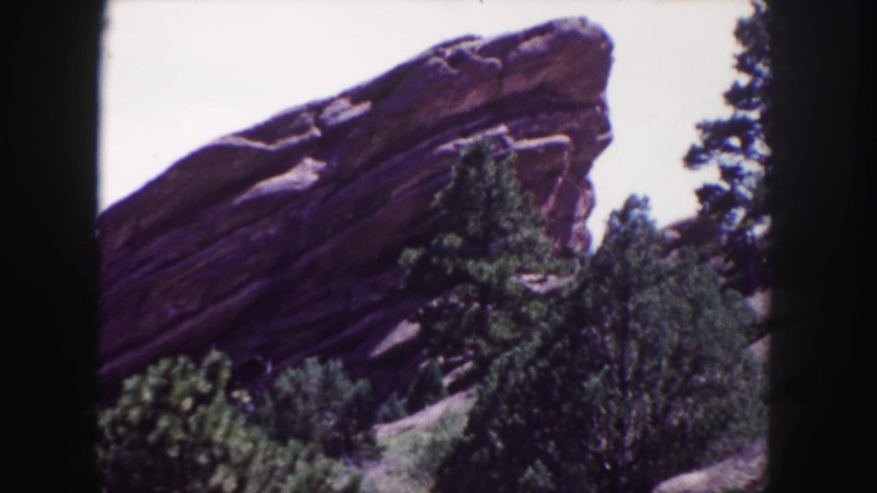 1958 8mm frame of red rock formations near Denver, with evergreen trees and scrub vegetation in the foreground.