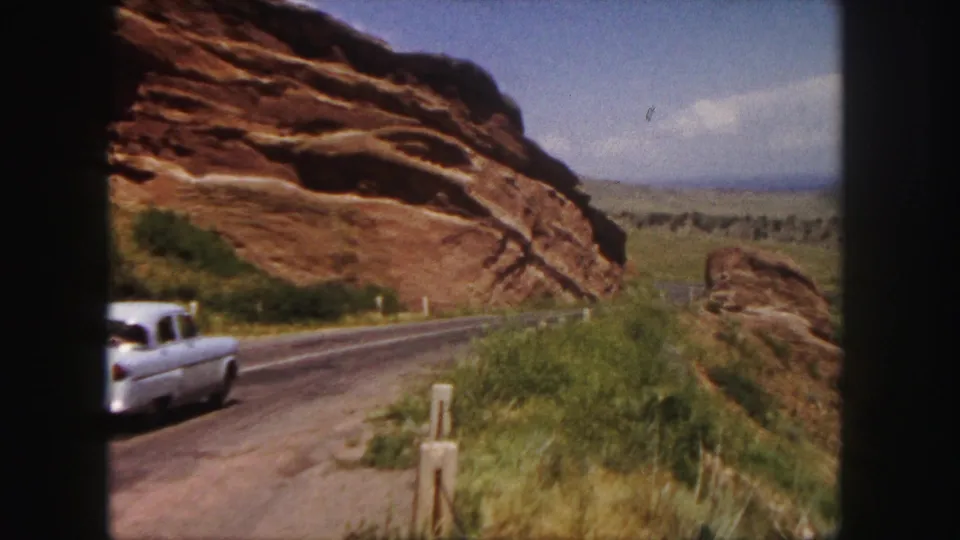 A white mid-century sedan passes along a rural road near Denver red rocks, with shrubs and roadside posts in view.