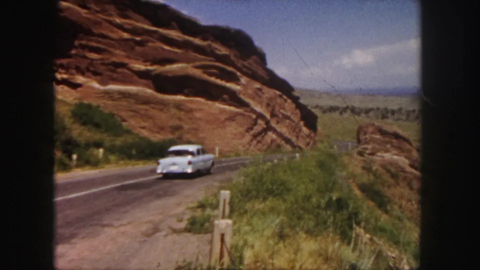 White sedan recedes on a paved road through Denver-area red rock formations, framed by brush, posts, and open sky.