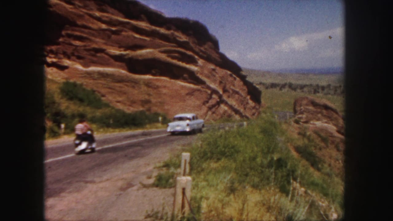White car and motorbike on a road beside a large red rock cliff.