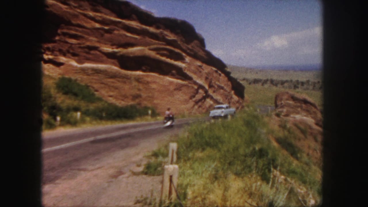 Motorbike and white car continue along the road beside the cliff.