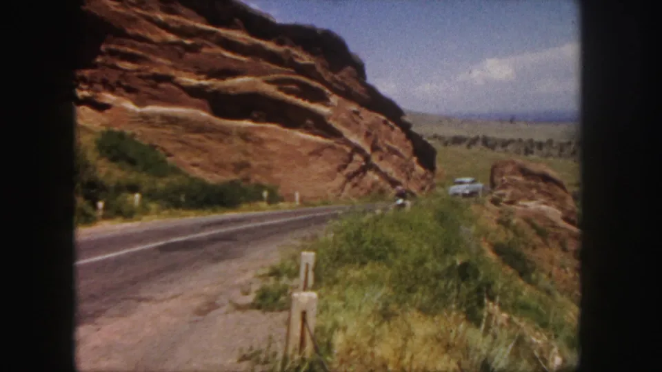 A distant car and motorbike travel away on a 1958 Denver-area road framed by red sandstone and scrub grass.
