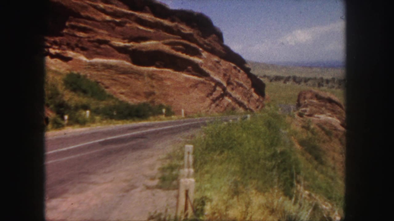 Paved road beside red rock cliffs with green roadside vegetation and blue sky.