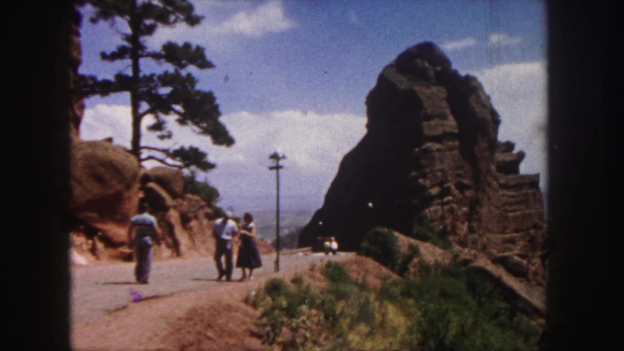 Visitors in a 1958 Denver-area mountain landscape walk near red rock outcrops beside a paved road.