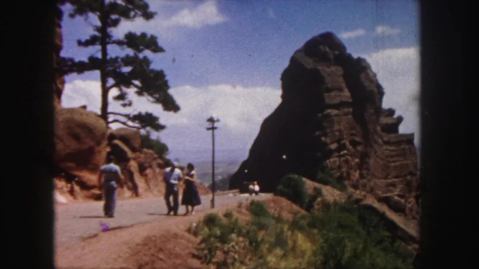 Visitors in a 1958 Denver-area mountain landscape walk near red rock outcrops beside a paved road.