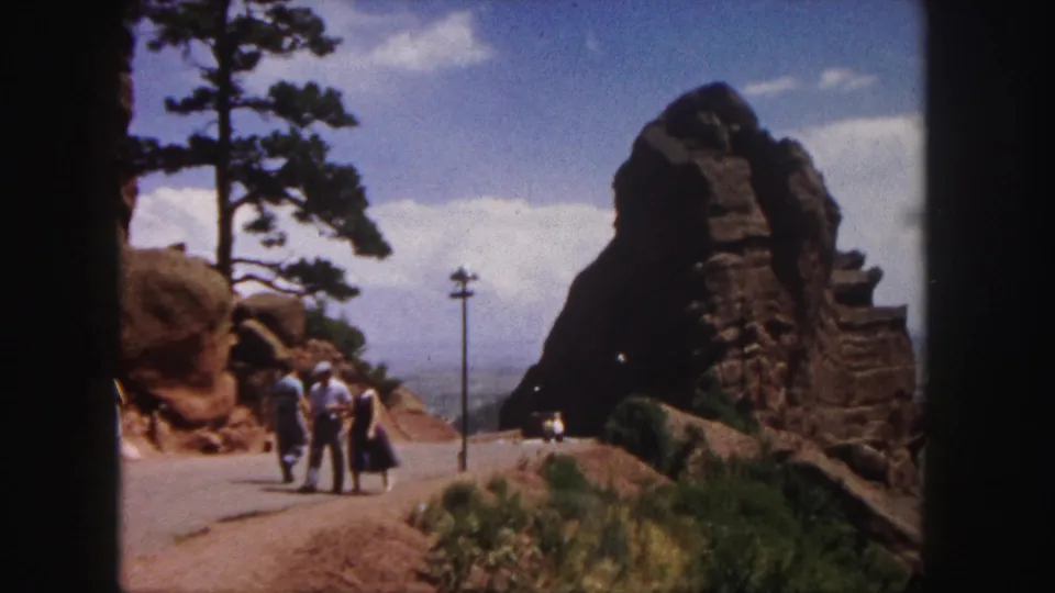 1958 home-movie frame of people walking along a red rock mountain road near Denver, with trees and a utility pole visible.