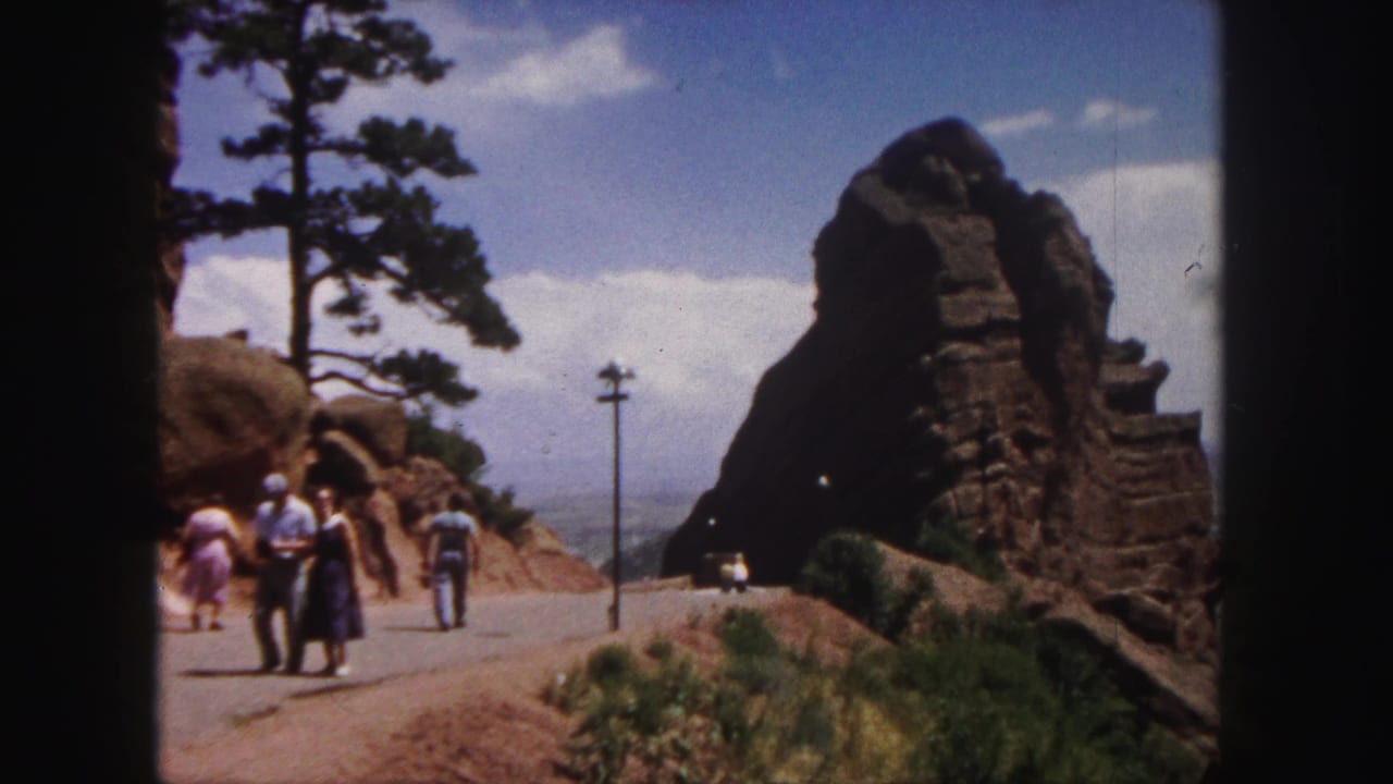 1958 Denver-area home-movie frame of visitors walking on a paved road beside red rock formations and a tall pine tree.