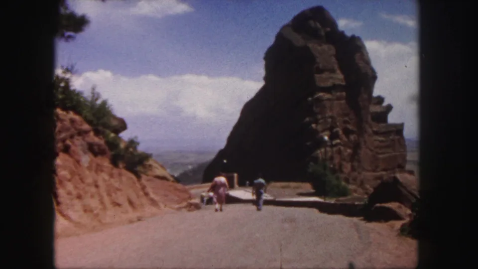 Handheld 8mm frame showing a road between red rock slopes, with visitors walking toward a large formation in Denver metadata context.