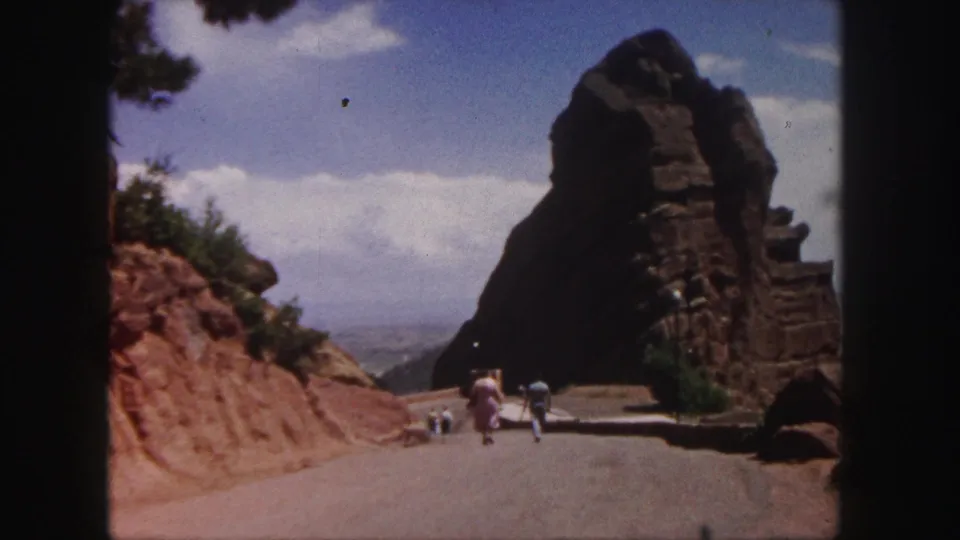 1958 Colorado travel frame with visitors receding along a paved road beside red rock cuts and a shadowed rock formation.
