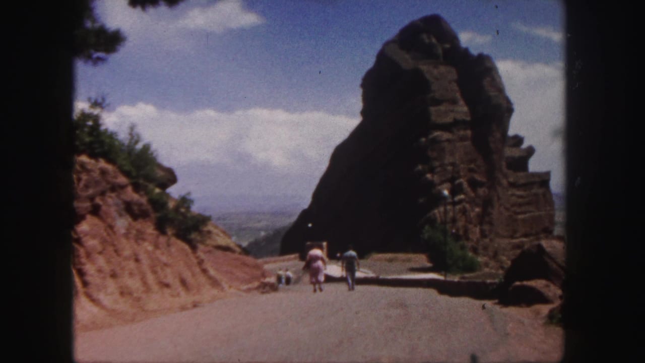 People walking along a road toward a large brown rock formation under a blue sky.