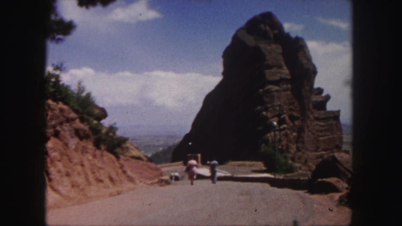 1958 Denver-area travel footage shows people on a road approaching a dark brown rock outcrop, with clouds and distant terrain beyond.