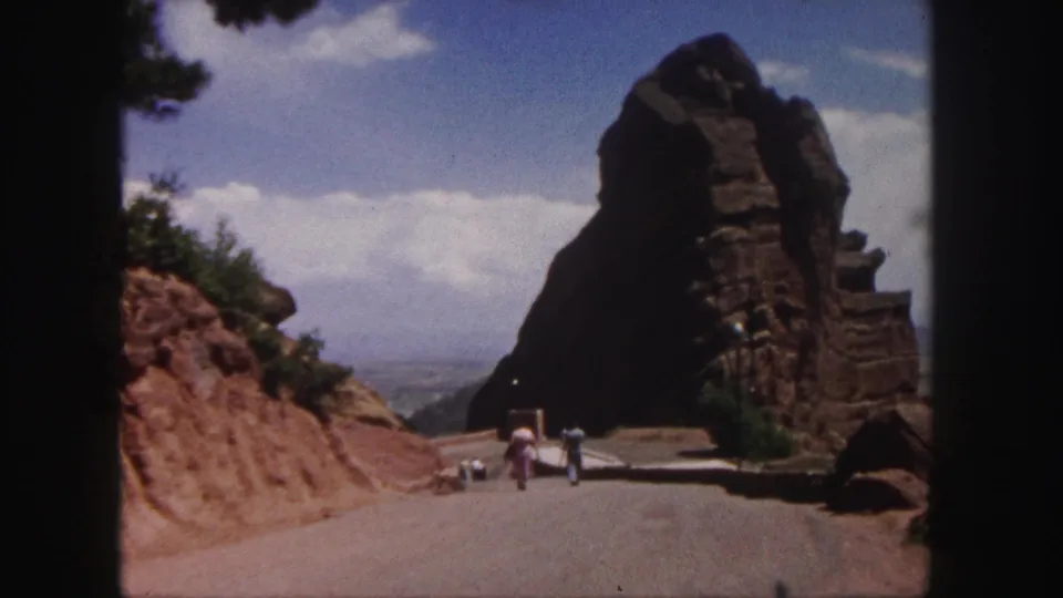 A 1958 8mm frame from Denver shows a visitor-shot road scene with people walking toward a tall brown rock formation.