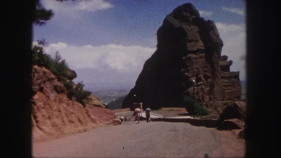 Final 1958 Denver-area frame shows visitors near the base of a large rock formation, with road, clouds, and distant landscape visible.