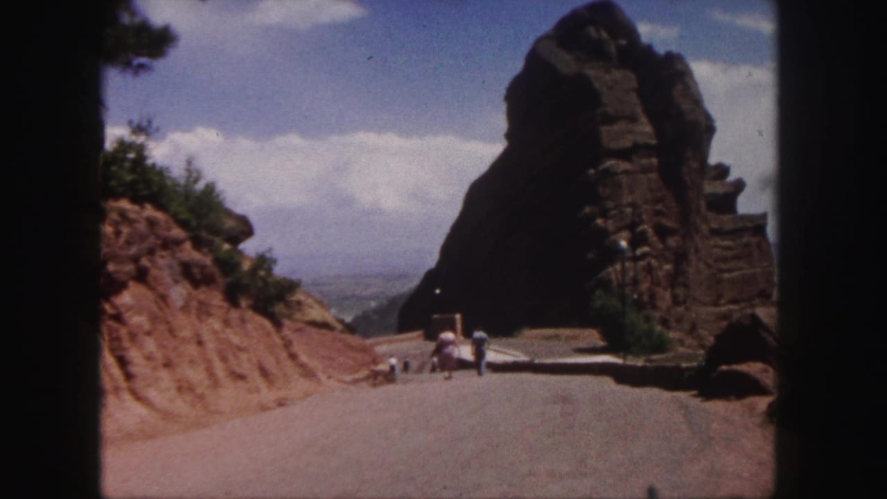 1958 home-movie frame of a road approaching Red Rocks Amphitheatre near Denver, with red rock walls and small visitors ahead.