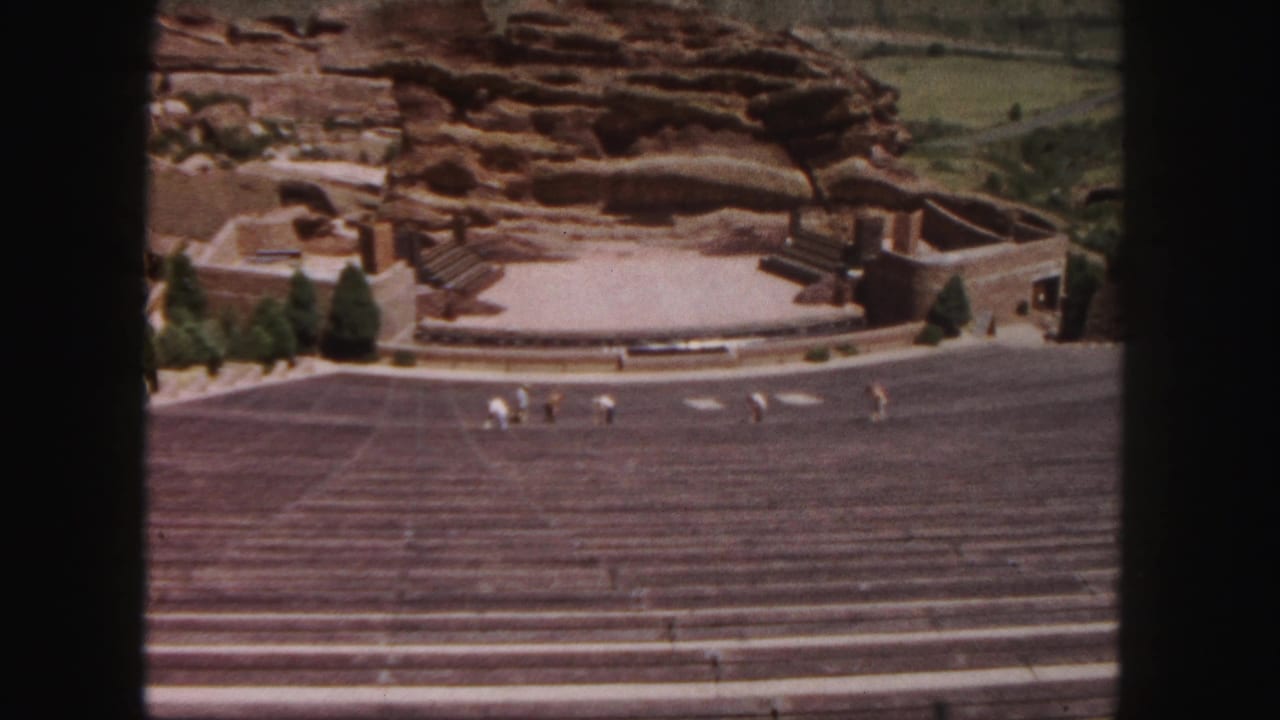 Wide amphitheatre seating with red rock backdrop and small visitors crossing the steps.
