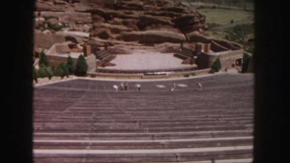Elevated 1958 home-movie view across Red Rocks Amphitheatre, showing rows of seating, the stage, trees, and small visitor figures.