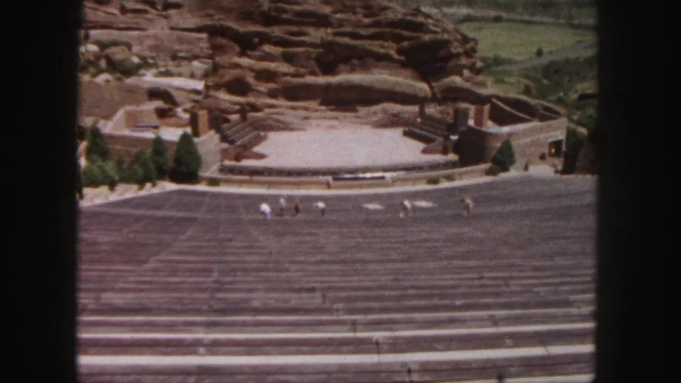 1958 wide shot of Red Rocks Amphitheatre near Denver, with stepped seating, stage structures, trees, and visitors moving across the site.