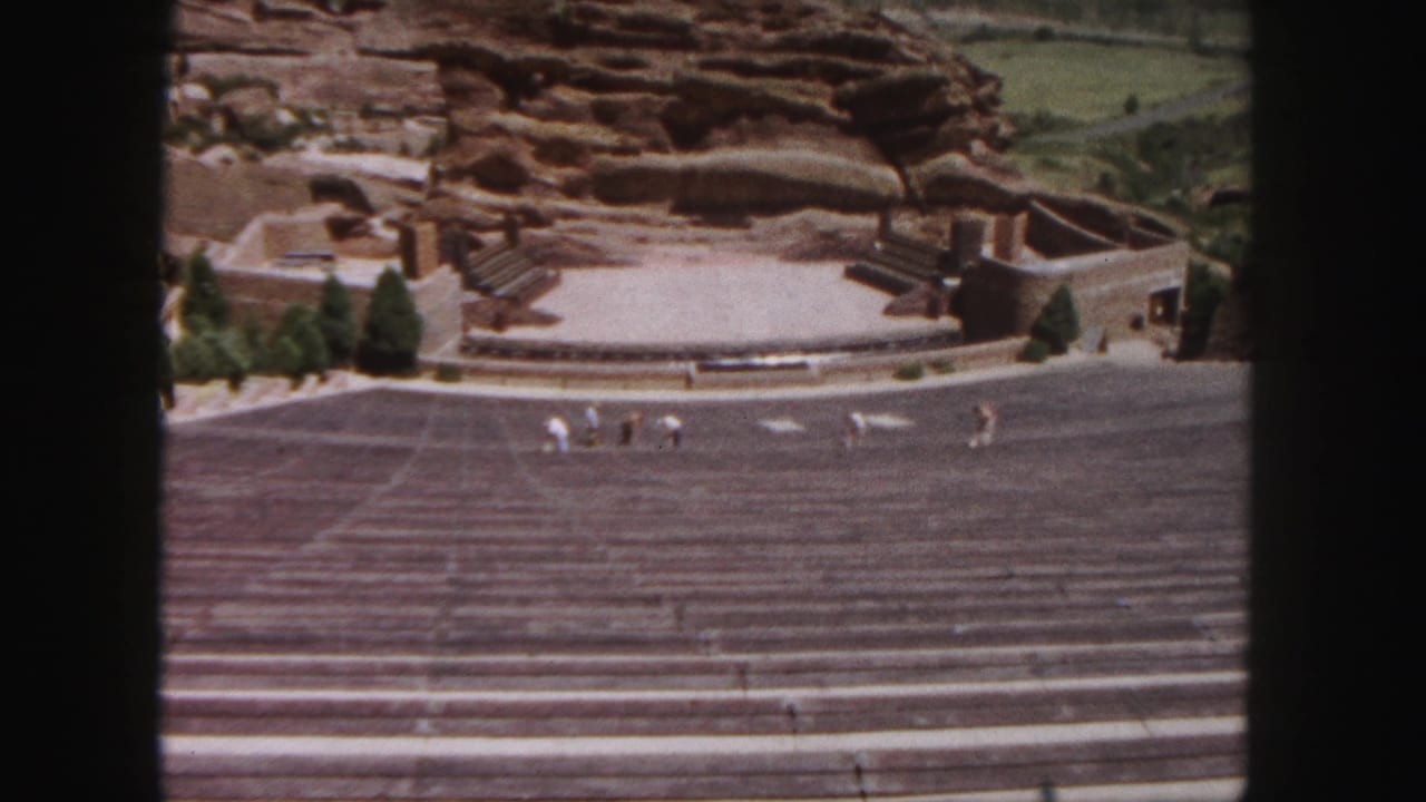 1958 8mm view of Red Rocks Amphitheatre in Denver, with tiered seating, open stage, red sandstone, and small groups of visitors.