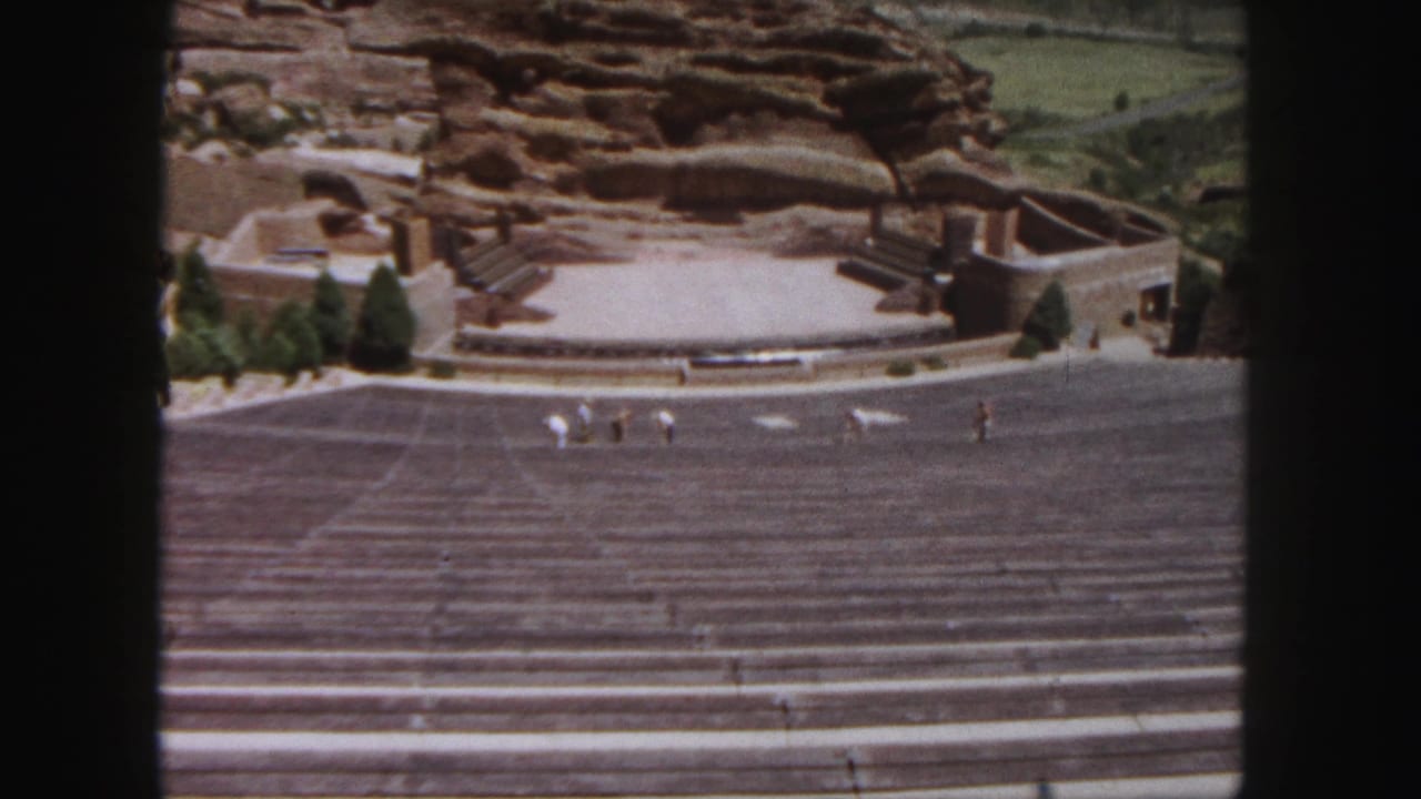 Wide amphitheatre view with people crossing the stage area below red rock formations.
