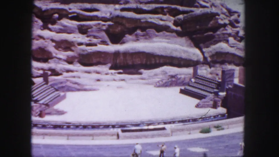The camera frames the Red Rocks stage and sandstone backdrop more tightly, with stairs, a roadway edge, and several visitors visible near the lower frame.