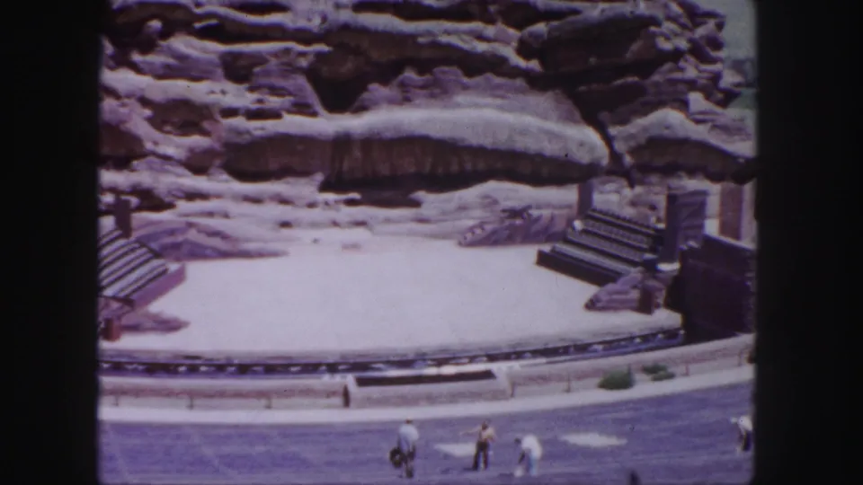 Late-frame view of Red Rocks Amphitheatre shows the pale stage, red sandstone wall, stair structures, roadway, and visitors walking along the front edge.