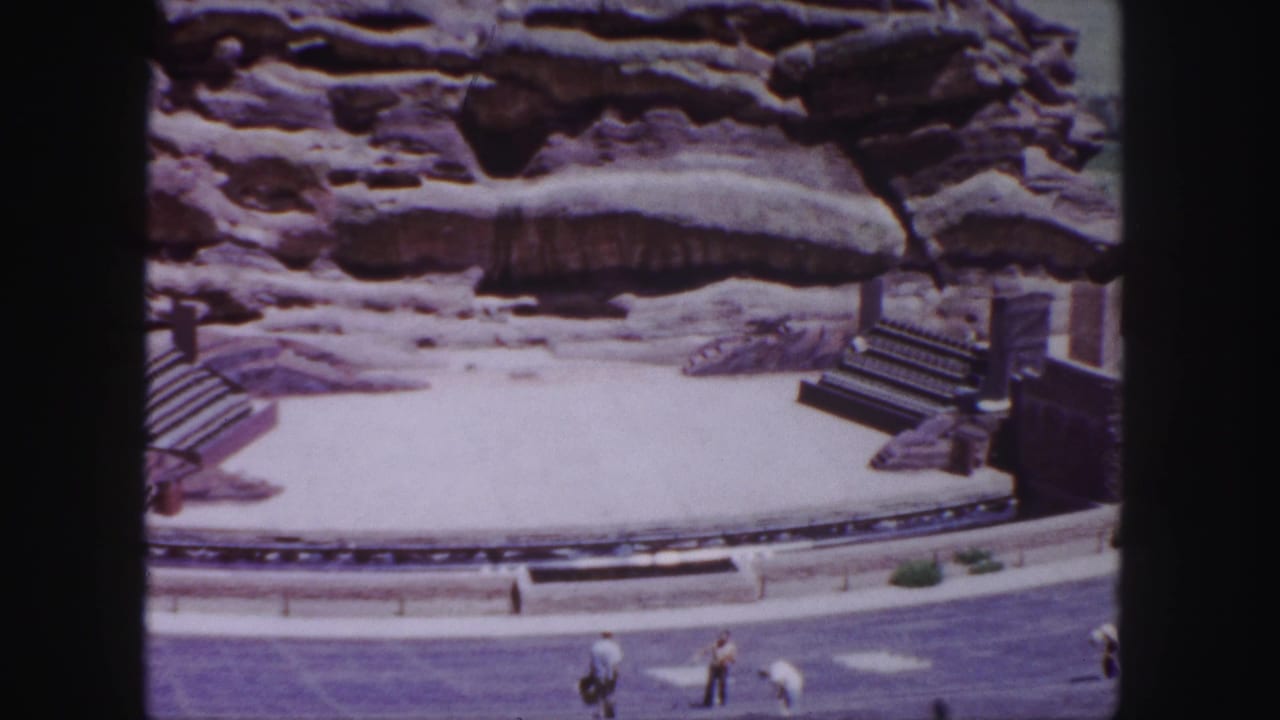 1958 Denver home-movie frame showing a circular amphitheater-like area with rock formations, tiered seating, stairs, and distant visitors.