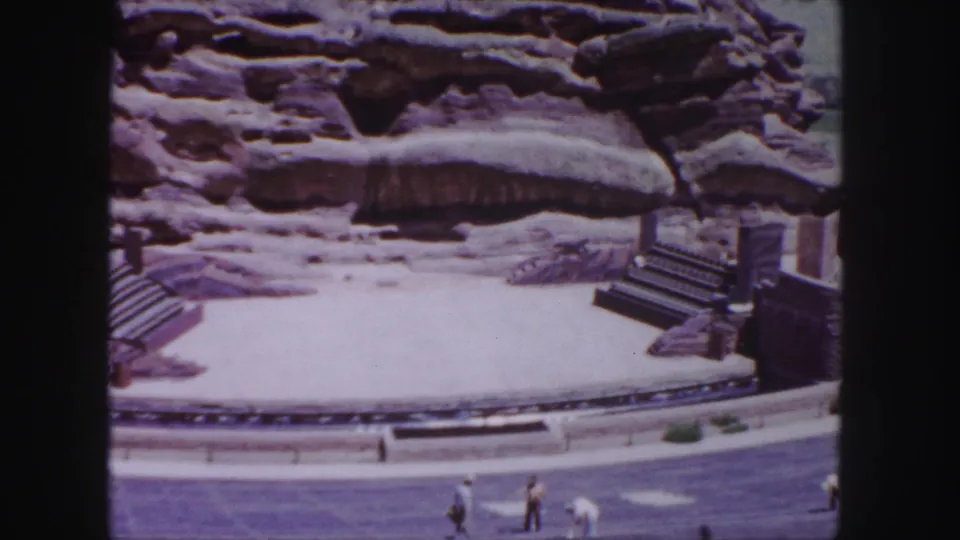 1958 Denver footage frame with an amphitheater-like stage, rocky backdrop, stairs, seating, and several visitors walking along the foreground.
